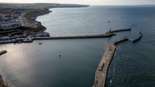 An aerial view of a harbor with breakwaters, a town on a cliffside, boats in the water, and lighthouses  in Whitby, UK.