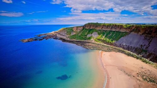 Aerial view of a scenic coastal landscape featuring a sandy beach, clear blue waters, and green cliffs under a partly cloudy sky.