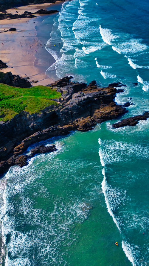 Aerial view of a beautiful coastline featuring sandy beaches, rocky cliffs, and turquoise waves crashing onto the shore. The landscape is lush with greenery, and the ocean displays a vibrant blue color, creating a picturesque scene.