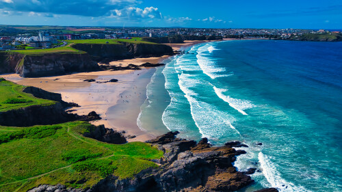 Aerial view of a beautiful beach with golden sand and turquoise waves. Lush green cliffs surround the beach, and a small town is visible in the background under a clear blue sky with scattered clouds.