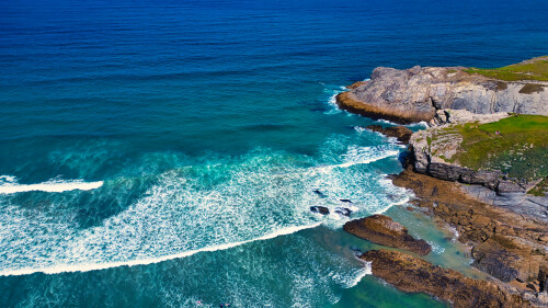 Aerial view of a rocky coastline with waves crashing against the shore. The vibrant blue ocean contrasts with the rugged rocks and green grass on the cliffs. The scene captures the beauty of nature and the dynamic movement of water.