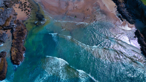 Aerial view of a beach with clear blue waters and sandy shores. Waves gently crash on the shore, creating a beautiful contrast with the rocky formations. People can be seen walking along the beach, enjoying the sunny day.