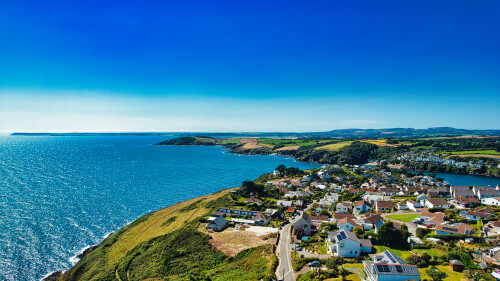 Aerial view of a coastal village with houses along the shoreline, surrounded by lush green fields and a sparkling blue sea under a clear sky. The landscape features a mix of residential areas and natural beauty, showcasing the harmony between human habitation and nature.