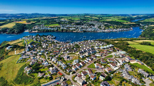 Aerial view of a picturesque coastal village surrounded by lush green hills and a river. The village features a mix of residential homes and boats in the water, with a clear blue sky overhead.