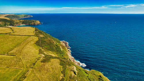 Aerial view of a coastal landscape featuring green fields and cliffs meeting the blue ocean. The horizon is clear with a few boats visible in the distance, showcasing a serene and picturesque natural setting.