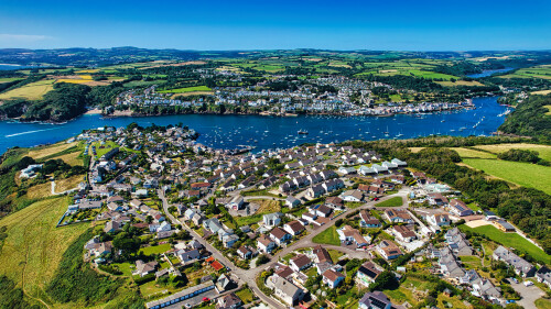 Aerial view of a picturesque coastal town with lush green hills, a river, and numerous boats in the water. The town features a mix of residential homes and scenic landscapes, showcasing a vibrant community surrounded by nature.