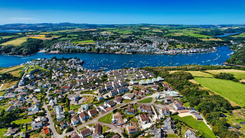 Aerial view of a picturesque coastal town with lush green hills, a river filled with boats, and residential areas. The landscape features a mix of houses and natural scenery, showcasing the beauty of the countryside and waterfront.