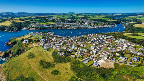 Aerial view of a picturesque coastal village surrounded by lush green hills and a serene bay filled with boats. The village features charming houses and a winding river, showcasing the beauty of nature and architecture.