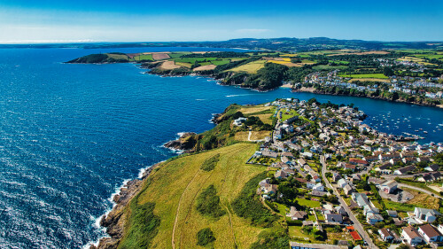 Aerial view of a coastal village with houses along the shoreline, lush green fields, and a clear blue sea. The landscape features rolling hills and a picturesque coastline, showcasing the beauty of nature and rural living.