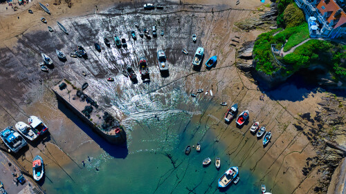 Aerial view of a harbor at low tide, showcasing numerous boats resting on the exposed mud. The water is calm with a mix of blue and green hues, and the shoreline features rocky formations and vegetation. The scene captures the tranquility of a coastal environment.