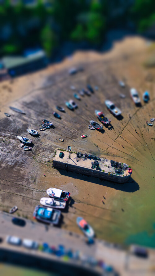 Aerial view of a beach with boats on the shore. The scene shows various boats parked on the sand during low tide, with some people walking around. The water is visible in the background, and greenery surrounds the area.