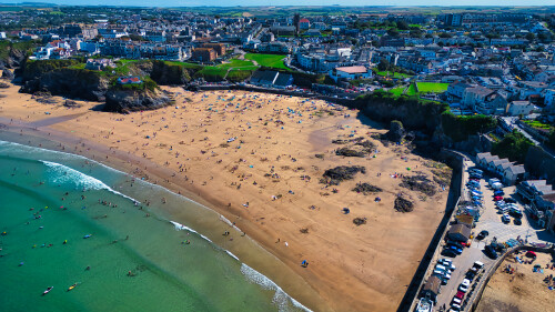 Aerial view of a sandy beach with people sunbathing and swimming, surrounded by cliffs and a coastal town. The beach is busy with umbrellas and towels, while the water is clear and inviting. The town features buildings and greenery in the background.