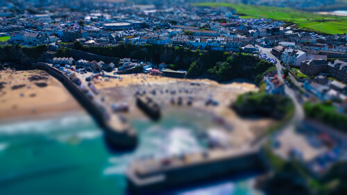 Aerial view of a coastal town with sandy beach, rocky shoreline, and colorful houses. The scene captures beachgoers enjoying the sun, with boats in the water and green fields in the background.