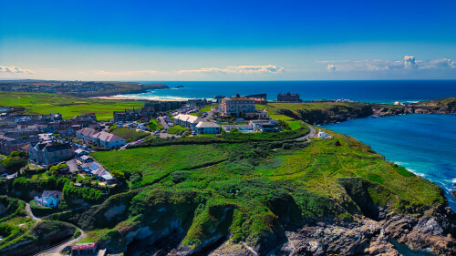 Aerial view of a coastal town with lush green hills, a hotel, and the ocean in the background. The sky is clear and blue, with a few clouds. The landscape features a mix of buildings, roads, and natural scenery, showcasing the beauty of the coastline.