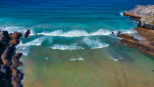 Aerial view of a beautiful beach with turquoise waters, gentle waves, and rocky formations. The shoreline features a mix of sand and rocks, with a few surfers in the water. The scene captures the tranquility and natural beauty of the coastal landscape.