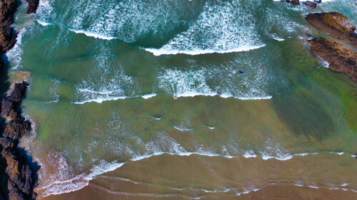 Aerial view of a beach with waves crashing onto the shore. The water is a mix of turquoise and green, with white foam from the waves. The sandy beach is visible, along with rocky formations on the sides.