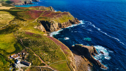 Aerial view of a rugged coastline with cliffs and vibrant green and purple vegetation. The blue ocean waves crash against the rocky shore, creating a picturesque landscape. A small settlement with white buildings is visible near the coast.