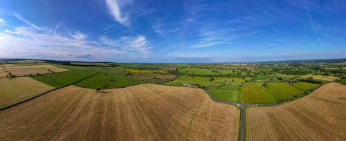 Panoramic aerial view of lush countryside with fields and a road under a blue sky.