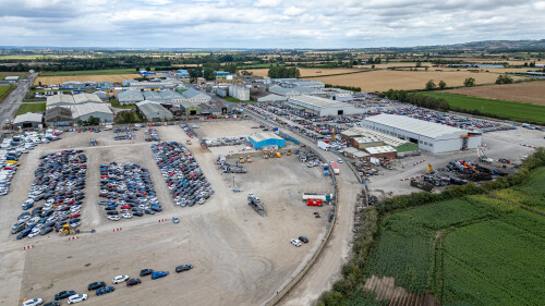 Aerial view of a car storage and processing facility, featuring large car parks, industrial buildings, and surrounding agricultural fields under a cloudy sky  at Copart yard in York, UK.