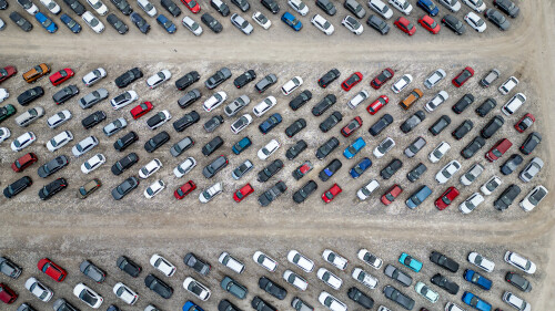 An overhead aerial view shows a large outdoor parking lot filled with numerous vehicles. The cars are arranged in neat rows and are predominantly gray, white, and black, with a few red and blue accents  at Copart yard in York, UK.