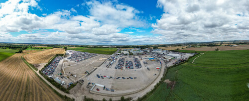 Aerial panoramic view of a large car scrapyard and surrounding farmland under a partly cloudy blue sky  at Copart yard in York, UK.