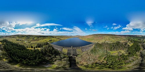 A panoramic view of a dam and reservoir surrounded by lush greenery and rolling hills under a bright blue sky at Scar House Reservoir, UK.