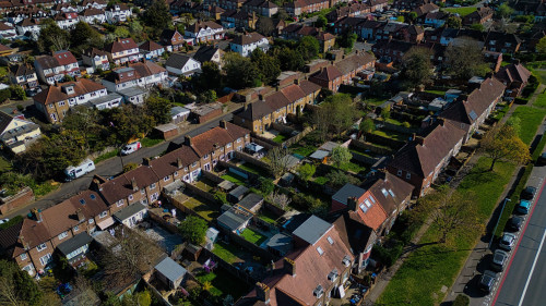Aerial view of a suburban neighborhood featuring rows of houses with gardens, trees, and parked cars on the street in Morden, London, UK.