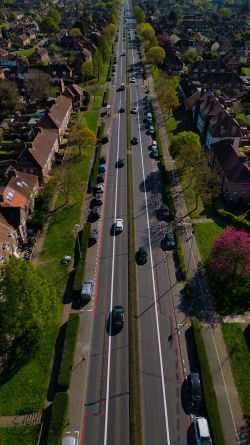 Aerial view of a straight road dividing residential area, lined with parked cars, trees, and houses on both sides in Morden, London, UK.
