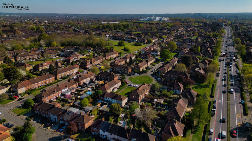 Aerial view of a suburban neighborhood with houses, trees, and a multi-lane road with traffic, under a clear blue sky in Morden, London, UK.