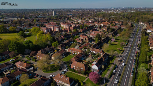 Aerial shot of a suburban neighborhood with houses, trees, a park, and a multi-lane road. City skyline in the distance in Morden, London, UK.