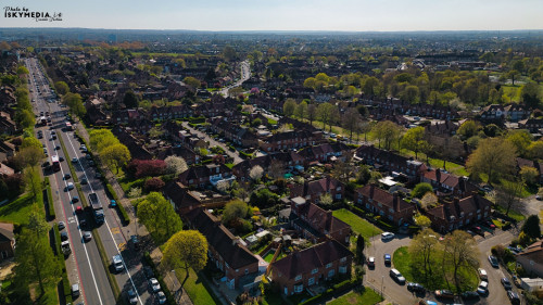 Aerial view of a suburban neighborhood with houses, roads, trees, and cars under a blue sky in Morden, London, UK.