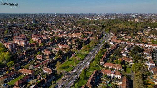 Aerial view of a suburban landscape featuring houses, a road, and green spaces under a clear blue sky in Morden, London, UK.