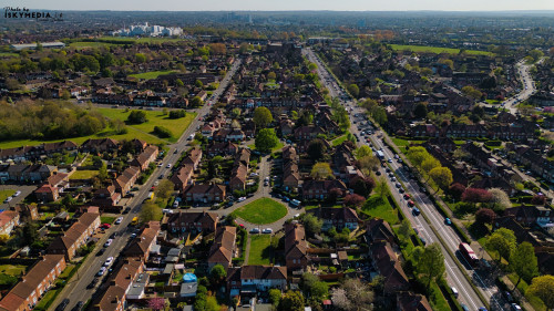 Aerial view of a suburban neighborhood with streets, houses, cars, and green spaces, taken during the day in Morden, London, UK.