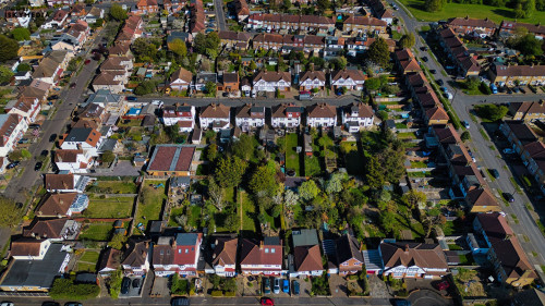 Aerial view of a suburban neighborhood with rows of houses, green lawns, trees, and streets with parked cars in Morden, London, UK.