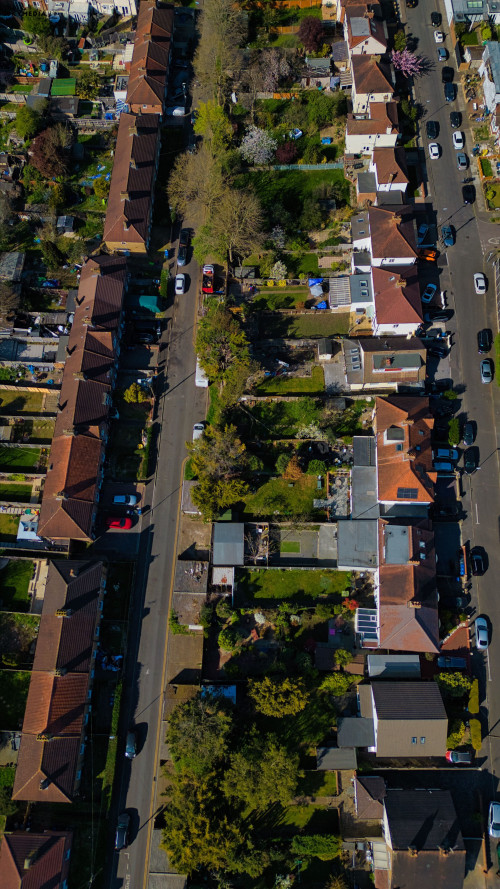 Aerial view of a residential street lined with houses, trees, and parked cars on a sunny day in Morden, London, UK.