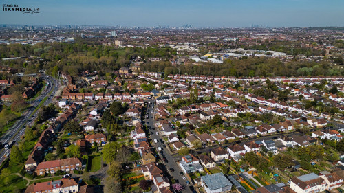 Aerial view of a dense suburban area with houses, roads, trees, and a distant cityscape under a clear blue sky in Morden, London, UK.