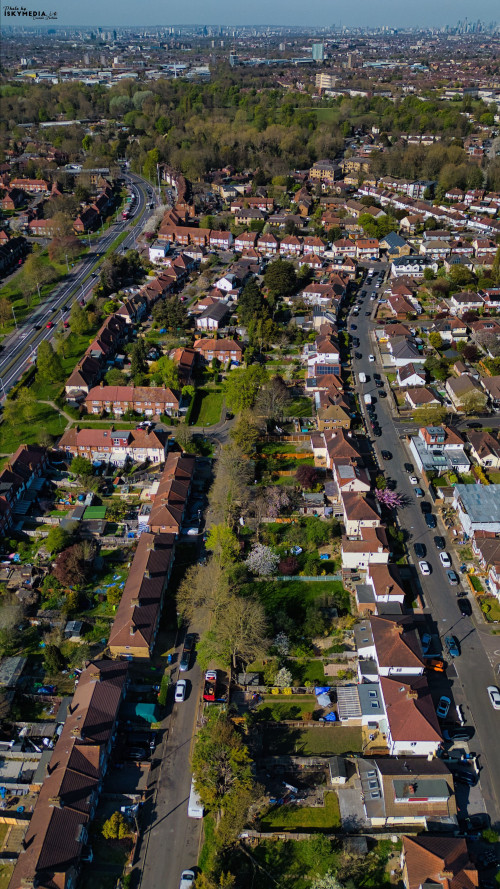 Aerial view of a suburban neighborhood with houses, gardens, roads, and distant city skyline under a blue sky in Morden, London, UK.