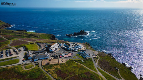 Aerial view of a coastal village with buildings near the shore, surrounded by green hills and a blue ocean. The scene captures the beauty of nature and human settlement by the sea, with a parking area and pathways visible.