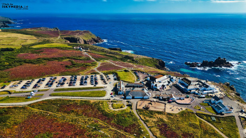 Aerial view of a coastal area featuring a parking lot, buildings, and rocky coastline. The landscape is lush with greenery and colorful vegetation, contrasting with the deep blue ocean. Clear skies enhance the scenic beauty.