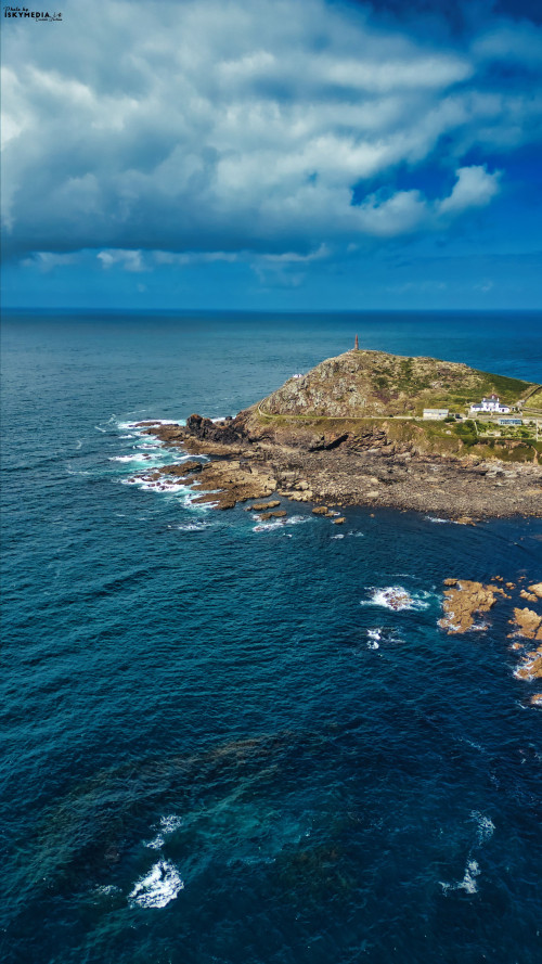 Aerial view of a rocky coastline with a small island. The island features a lighthouse on top, surrounded by deep blue ocean waters and dramatic clouds in the sky. The scene captures the beauty of nature and coastal landscapes.
