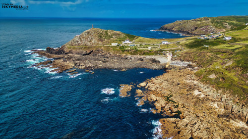 Aerial view of a coastal landscape featuring rocky shores, clear blue waters, and a green hillside. A small settlement with white buildings is visible near the shore, along with a lighthouse on the hilltop. The sky is bright with a few clouds.