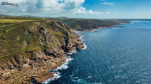 A stunning aerial view of rugged cliffs along a coastline, with clear blue waters and a bright sky. The landscape features green hills and rocky shorelines, showcasing the natural beauty of the area.