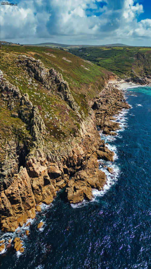Aerial view of rugged coastline with rocky cliffs and clear blue ocean. The landscape features green hills in the background under a partly cloudy sky, showcasing the natural beauty of the coastal area.