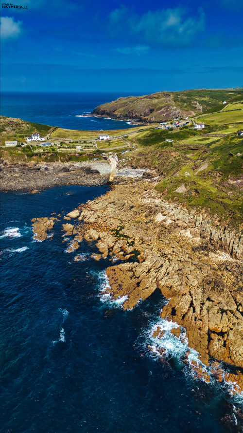 Aerial view of rugged coastline with rocky cliffs and blue ocean. Green hills in the background and a few houses dotting the landscape. The scene captures the beauty of nature and coastal geography.