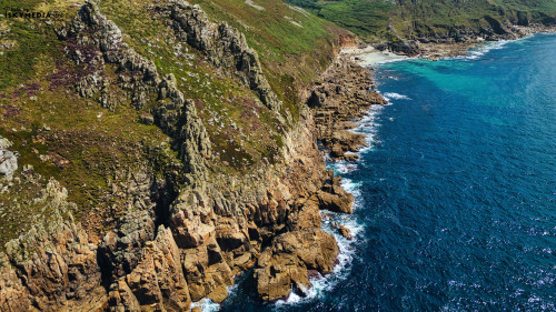 Aerial view of a rugged coastline with rocky cliffs and clear blue waters. The landscape features green hills in the background and waves crashing against the rocks, showcasing the natural beauty of the coastal environment.