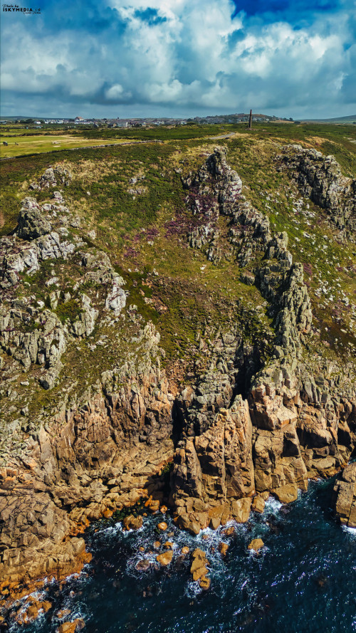 Aerial view of rugged coastal cliffs with rocky formations and waves crashing against them. The landscape features green hills and a cloudy sky in the background, showcasing the natural beauty of the coastline.