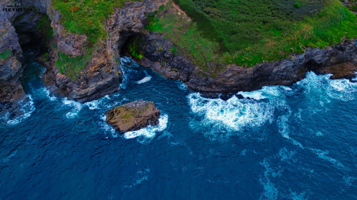 Aerial view of a rocky coastline with waves crashing against the shore. The vibrant blue ocean contrasts with the rugged cliffs and green vegetation on the land. A small rock formation is visible in the water, surrounded by foamy waves.