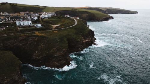 A scenic coastal view featuring a small village perched on a cliff overlooking the ocean. The waves crash against the rocky shore, with lush green hills in the background under a cloudy sky.
