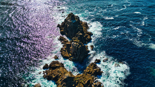 Aerial view of rocky formations surrounded by vibrant blue ocean waves. The sunlight reflects off the water, creating a shimmering effect. The rugged coastline features various textures and colors, highlighting the natural beauty of the seascape.