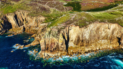 Aerial view of rugged cliffs along a coastline, with turquoise waters crashing against the rocks. The landscape features green vegetation on the cliffs and rocky formations, showcasing the natural beauty of the area.